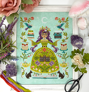 A cross stitch pattern featuring a stylized illustration of a woman surrounded by flowers and decorative elements, framed with flowers and scissors.