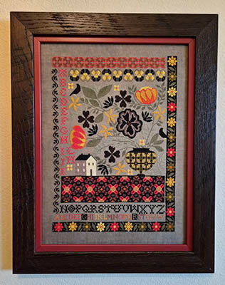 Framed cross-stitched sampler with floral and geometric patterns on a gray background.
