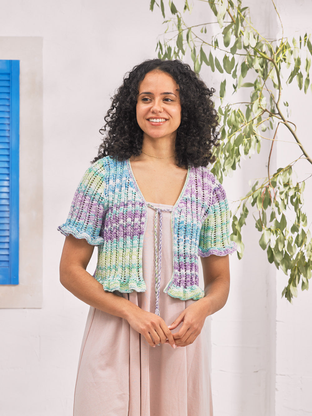 Woman wearing a colorful knitted cardigan standing in a room with plants and a painting.