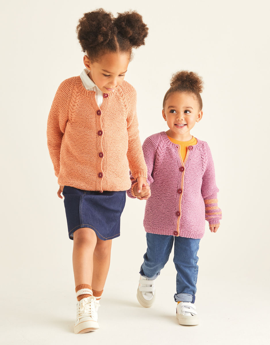 Two children wearing knitted raglan style cardigans with a cable detail, holding hands on a plain background
