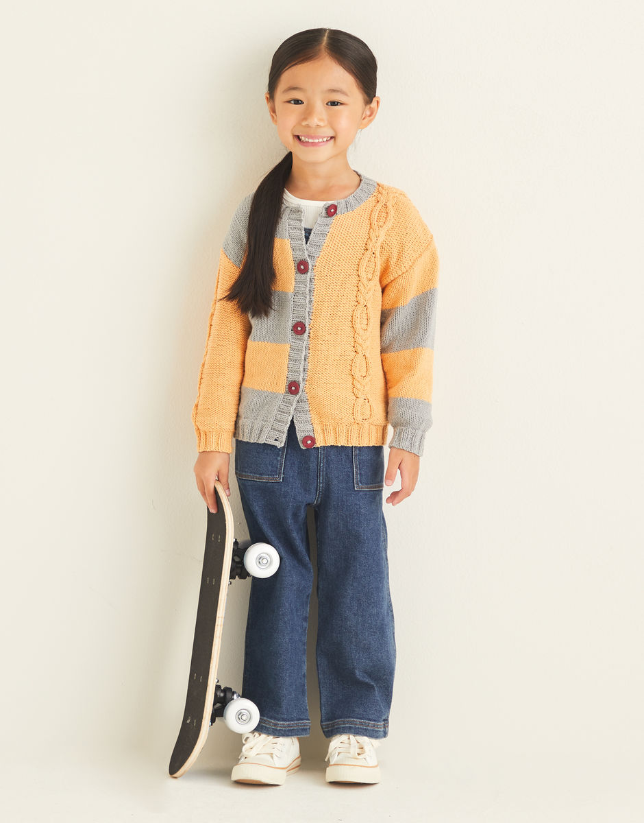 Child wearing a yellow cardigan with a striped section, and a cable detail,  holding a skateboard on a plain background