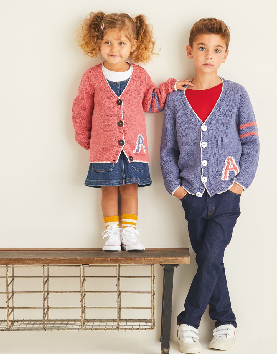 Two children wearing Varsity style cardigans with a letter 'A' design, standing on a bench against a plain background.