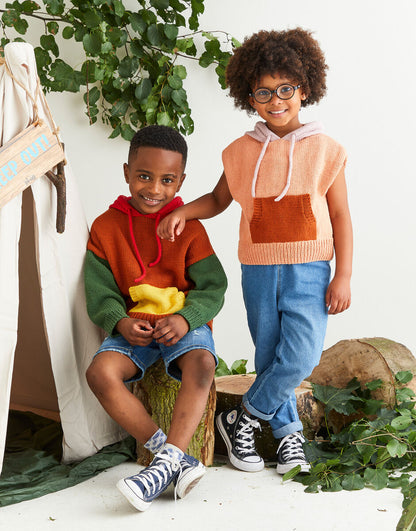 Two children wearing colorful hoodies and jeans standing in a natural setting with plants.