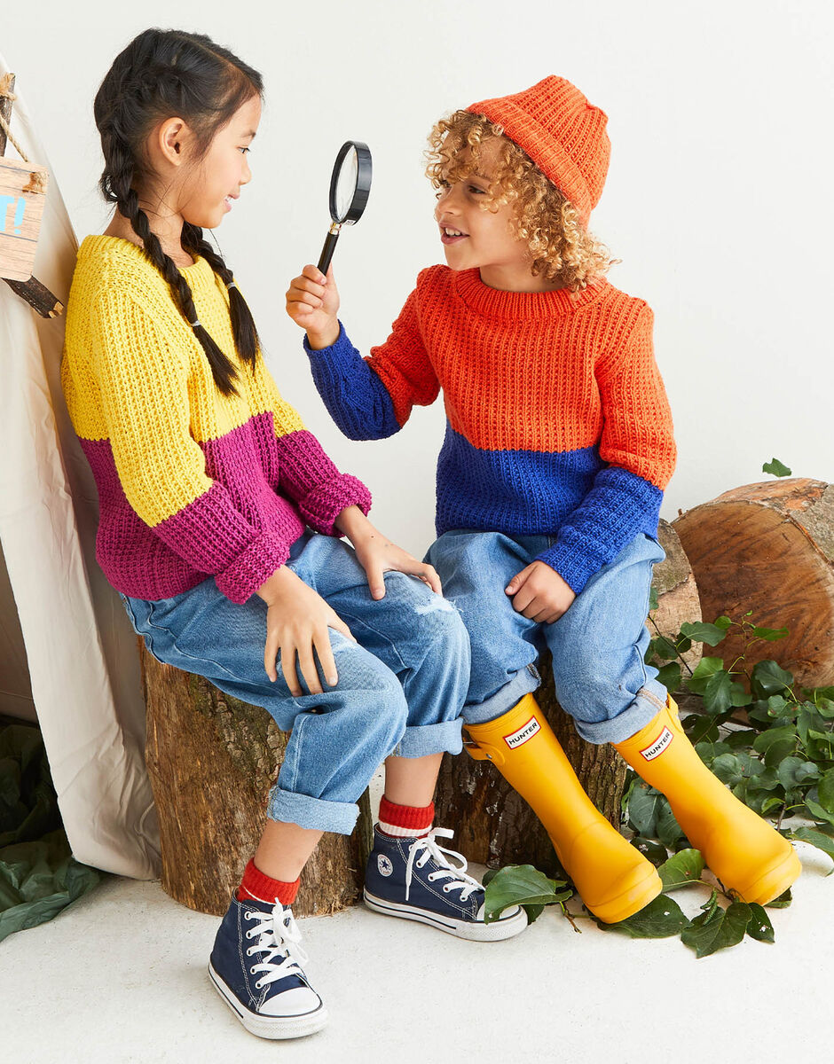 Two children sitting on logs wearing colorful sweaters and rain boots, one holding a magnifying glass.