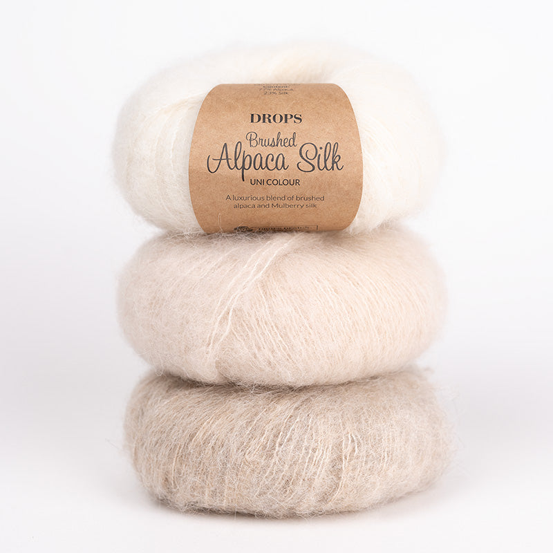 Three stacked skeins of DROPS Brushed Alpaca Silk yarn in a soft unicolor blend, with a kraft paper label on the top skein, photographed on a clean white background.